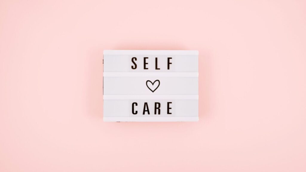 A white letterboard with the words 'Self Care' and a heart symbol, set against a soft pink background. Represents mindfulness, self-love, and mental wellness.